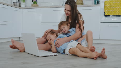 Mom-with-two-young-children-sitting-on-the-floor-in-a-modern-apartment-in-a-bright-interior-with-a-laptop.-Watch-the-laptop-screen-and-watch-family-photos.-Make-online-purchases-for-kids.